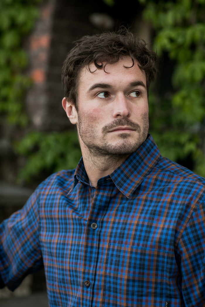 A man wears the Pladra Men's Tailwinds Shirt in Lagoon Blue, standing outdoors with greenery behind him.