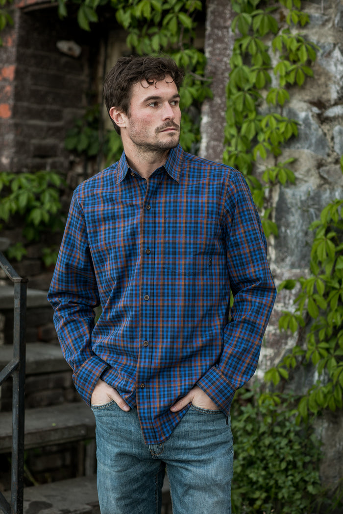 Man wears Pladra Men's Tailwinds Shirt in Lagoon Blue with jeans, standing outdoors by stone steps and vine-covered wall.