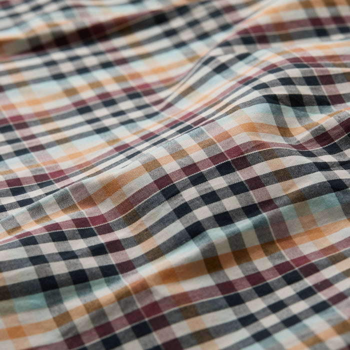 Close-up of the Men's Tailwinds Shirt in Pebble Brown, featuring a lightweight poplin weave. The soft cotton fabric displays gentle folds and shadows, highlighting its breathable comfort and plaid design.