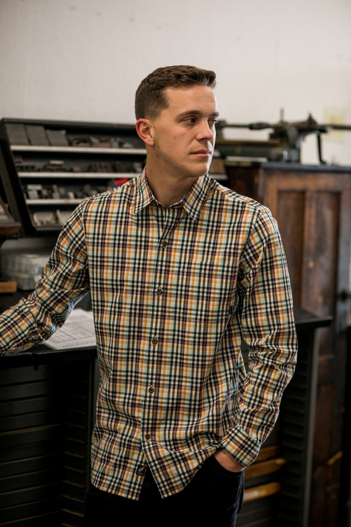 A man stands indoors with one hand in his pocket, wearing the Men's Tailwinds Shirt in Pebble Brown—a breathable cotton shirt with a plaid pattern. Shelves and cabinets are visible in the background as he looks to his left.