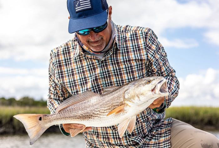 A man in sunglasses, a Men's Tailwinds Shirt–Pebble Brown, and a blue cap holds a large fish outdoors with grassy wetlands and a cloudy sky behind him.