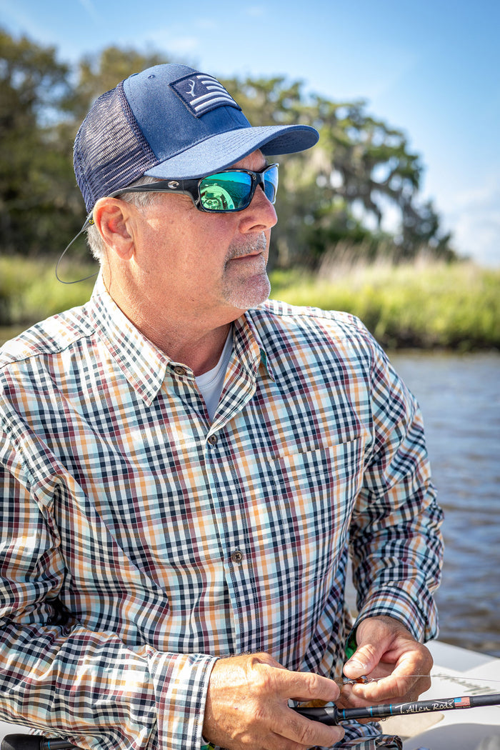 A man in a Men's Tailwinds Shirt- Pebble Brown, blue cap, and sunglasses sits by the water with fishing gear. He gazes to the side, surrounded by trees and grass on a sunny day.