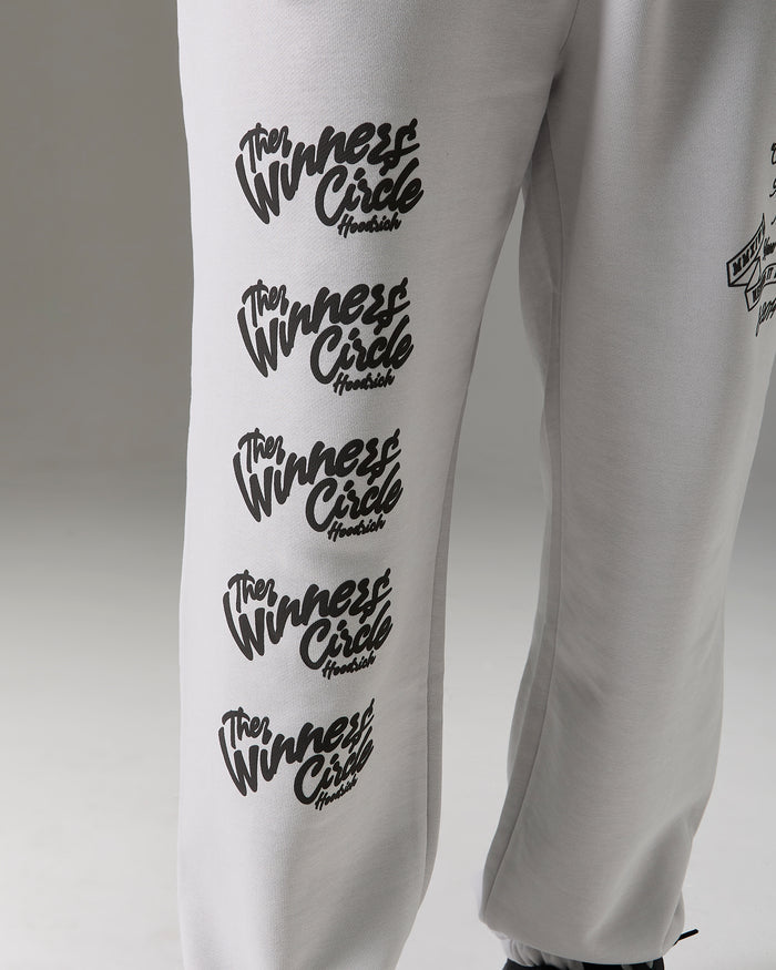 White sweatpants featuring a vertical black graphic print of the 'Winners Circle' logo repeated five times on the left leg, shown against a neutral gray background.