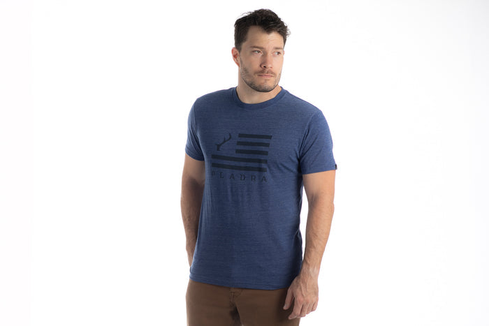 A man wears the Men's Tri-Blend Flag Graphic Tee in Beacon Blue Heather and brown pants, standing against a white background with one hand in his pocket—ideal for an outdoor lifestyle.