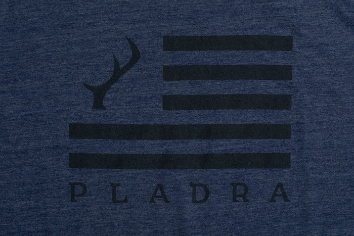 The Men's Tri-Blend Flag Graphic Tee in Beacon Blue Heather features a black flag with four stripes and an antler on the chest, plus PLADRA below, capturing an outdoor lifestyle.