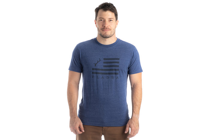 A man faces forward in a Men's Tri-Blend Flag Graphic Tee - Beacon Blue Heather, featuring a black flag graphic and PLADRA text on the front, paired with brown pants against a white background.