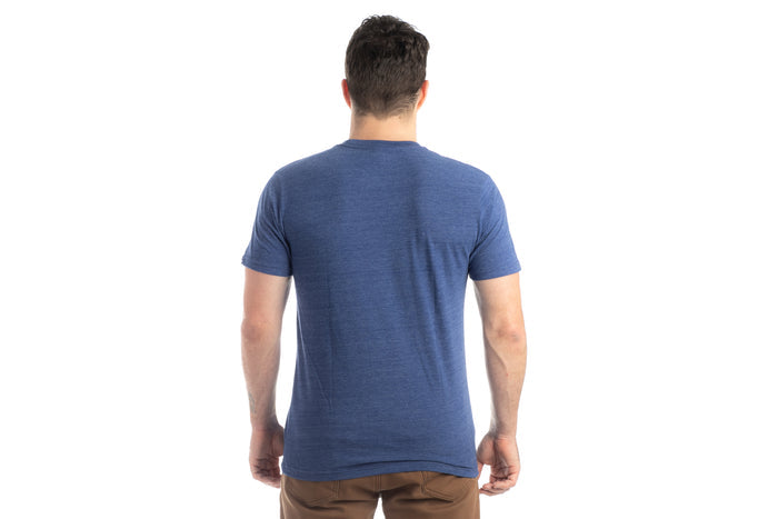 A person with short dark hair, wearing the Men's Tri-Blend Flag Graphic Tee in Beacon Blue Heather and brown pants, is standing facing away from the camera against a plain white background.