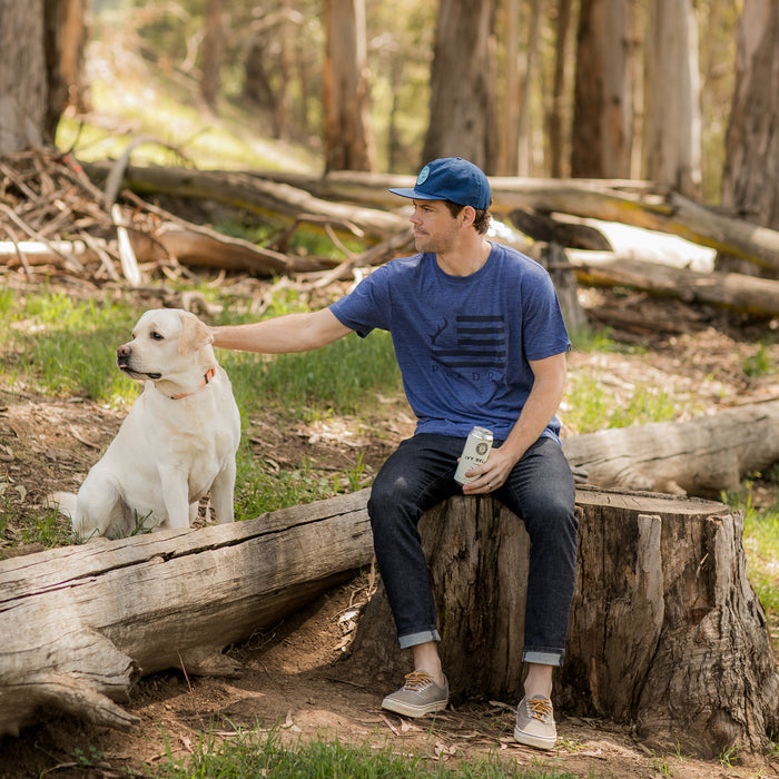 A man in a Men's Tri-Blend Flag Graphic Tee in Beacon Blue Heather and a cap sits on a tree stump, holding a drink can and petting a yellow Labrador. Sunlight streams through the trees, highlighting his relaxed outdoor lifestyle.