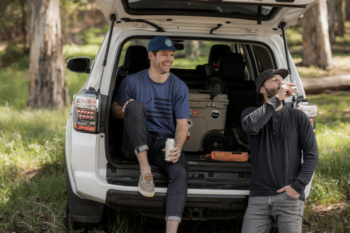 Two men enjoy the outdoors by an open SUV in the woods; one sits, holding a drink and smiling, while the other stands nearby, sipping from a can and wearing the Men's Tri-Blend Flag Graphic Tee in Beacon Blue Heather.