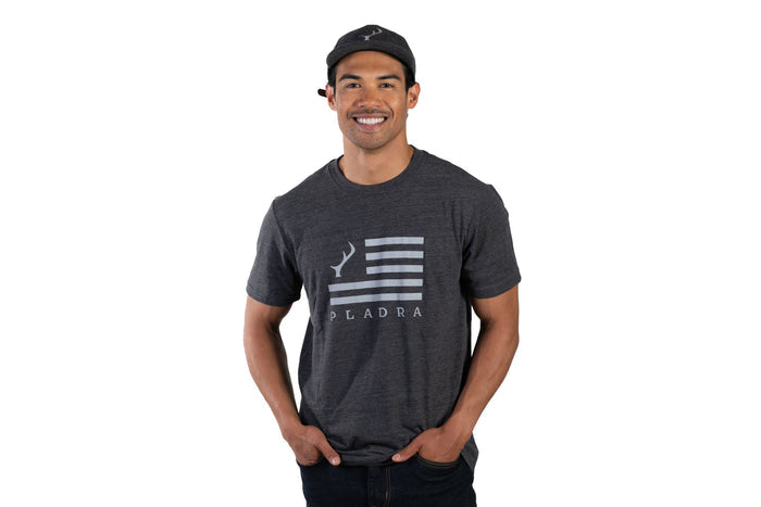 Man wearing Pladra Men's Tri-Blend Flag Graphic Tee in Charcoal Grey Heather and a hat, hands in pockets.