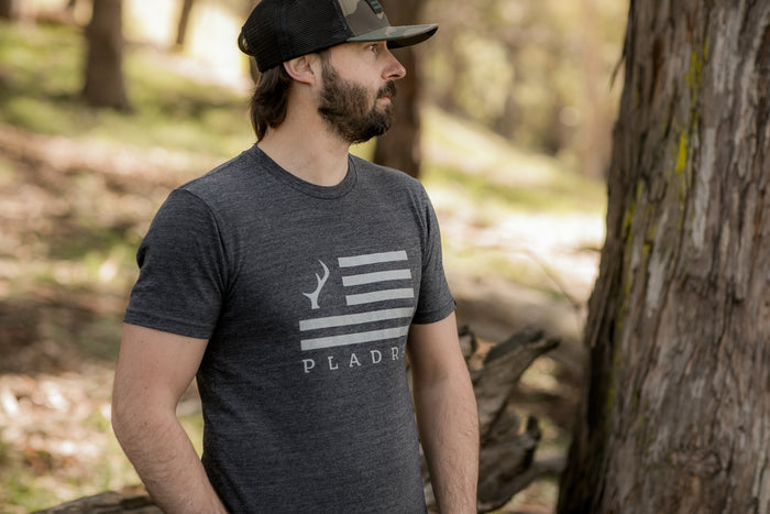 Man in a Pladra Men's Tri-Blend Flag Graphic Tee in Charcoal Grey Heather and black cap stands by a tree, looking aside.