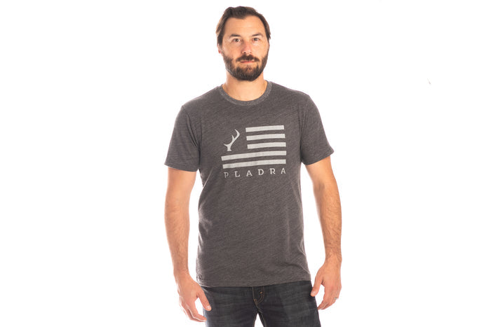 Man with a beard wears the Pladra Men's Tri-Blend Flag Graphic Tee in Charcoal Grey Heather and jeans against a white background.