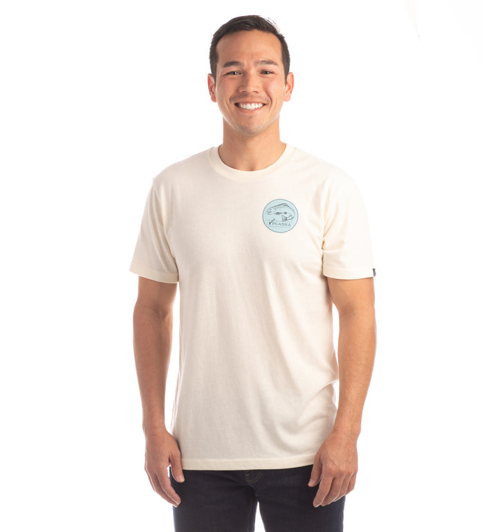 A man smiles while wearing the Men's Trout Tee- Tofu White Heather, a plain off-white jersey T-shirt with a small blue circular chest logo, paired with dark pants, against a white background.