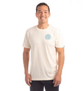 A man smiles while wearing the Men's Trout Tee- Tofu White Heather, a plain off-white jersey T-shirt with a small blue circular chest logo, paired with dark pants, against a white background.
