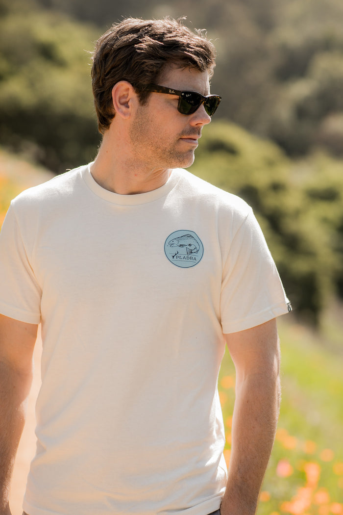 A man wearing the Men's Trout Tee in Tofu White Heather with a small blue logo and black sunglasses stands outdoors, looking to the side. The blurred background features greenery and orange flowers.