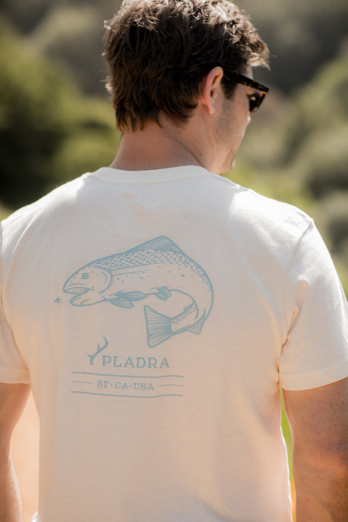 A man wearing sunglasses and a Men's Trout Tee- Tofu White Heather stands outdoors. The shirt’s back displays a blue trout graphic, PLADRA SF•CA•USA text, and a small antler graphic below.