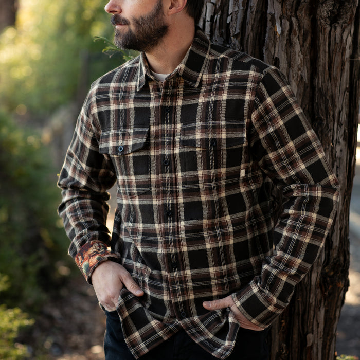A bearded man stands outdoors by a tree, wearing the Men's Workhorse Elite Flannel Shirt- Diamondback Brown with his hands in his pockets. The blurred background features greenery.