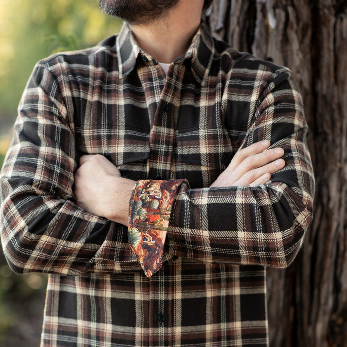 A bearded man stands outdoors with arms crossed, wearing the Men's Workhorse Elite Flannel Shirt in Diamondback Brown. The shirt's inner cuff reveals a colorful patterned fabric, and tree bark is visible in the background.