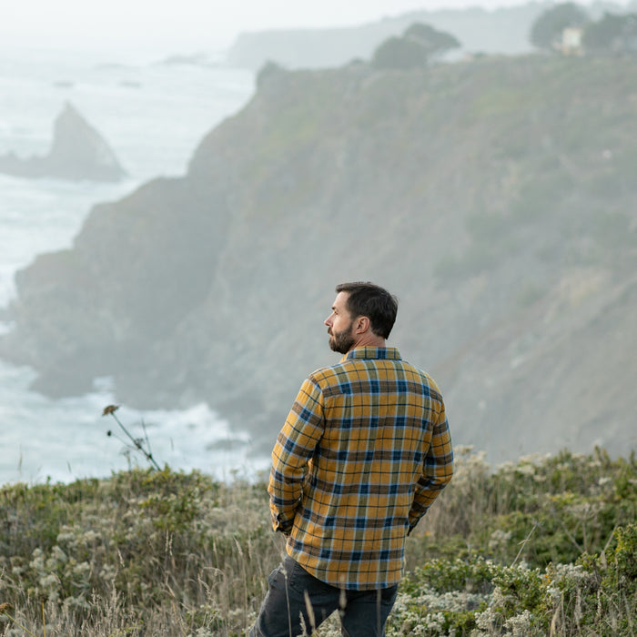 Man in Pladra Men's Workhorse Elite Flannel Shirt - Easton Gold stands on a cliff, looking out at the rocky coast.