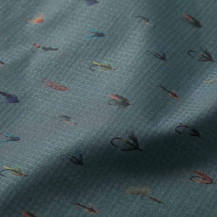 The Men's Yuba Fishing Shirt - Flies Print features quick-drying, UPF 50+ fabric with a subtle grid texture and a repeating pattern of small, colorful fishing flies on a muted blue-green background.