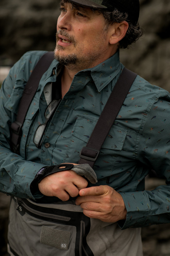 A man in the Men's Yuba Fishing Shirt—Flies Print, paired with waders, adjusts his chest straps. With a short beard, cap, and glasses hanging from his shirt, he enjoys this outdoor-ready top's quick-drying comfort by the water.