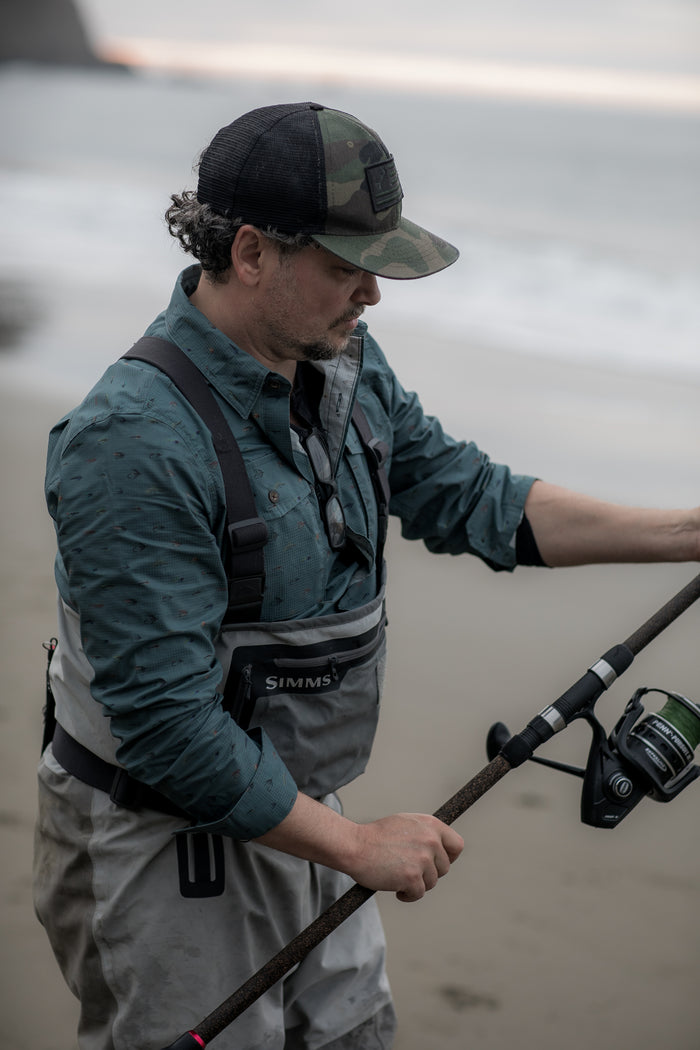 A man wearing a cap, sunglasses, and the Men's Yuba Fishing Shirt - Flies Print stands on a sandy beach with a fishing rod, the ocean in the background.
