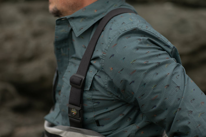 A person wearing the Men's Yuba Fishing Shirt - Flies Print in teal with black suspenders, standing outdoors near a rocky surface.