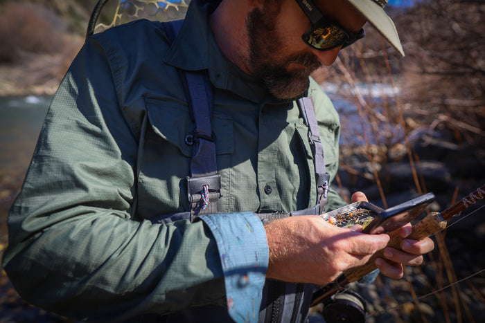 Man in Pladra Men's Yuba Fishing Shirt - Kombu Green, by the river with a rod, checks his tackle box.