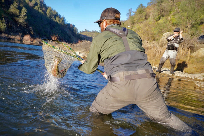 Two anglers fish; one nets a catch, the other sports Pladra’s Men's Yuba Fishing Shirt in Kombu Green with UPF 50+ protection.