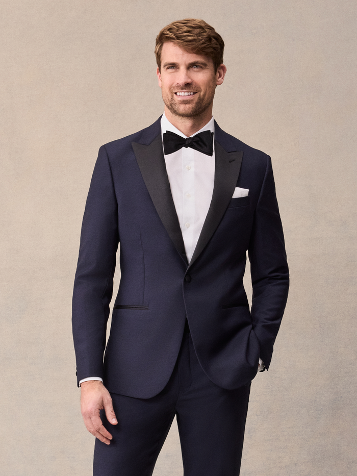 A man in a midnight blue tuxedo with black satin peak lapels, black bow tie, and white pocket square.