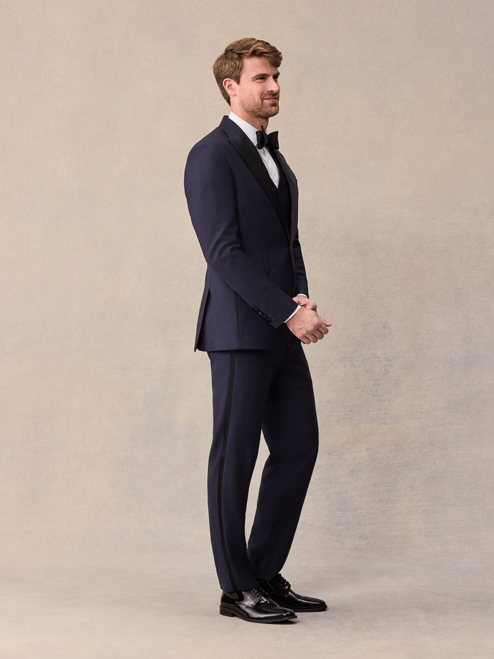 A man in a midnight blue tuxedo with black satin peak lapels, matching vest, side profile, full length.