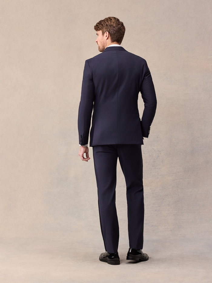 Back view of a man in a midnight blue tuxedo jacket and trousers, full length on a beige backdrop.