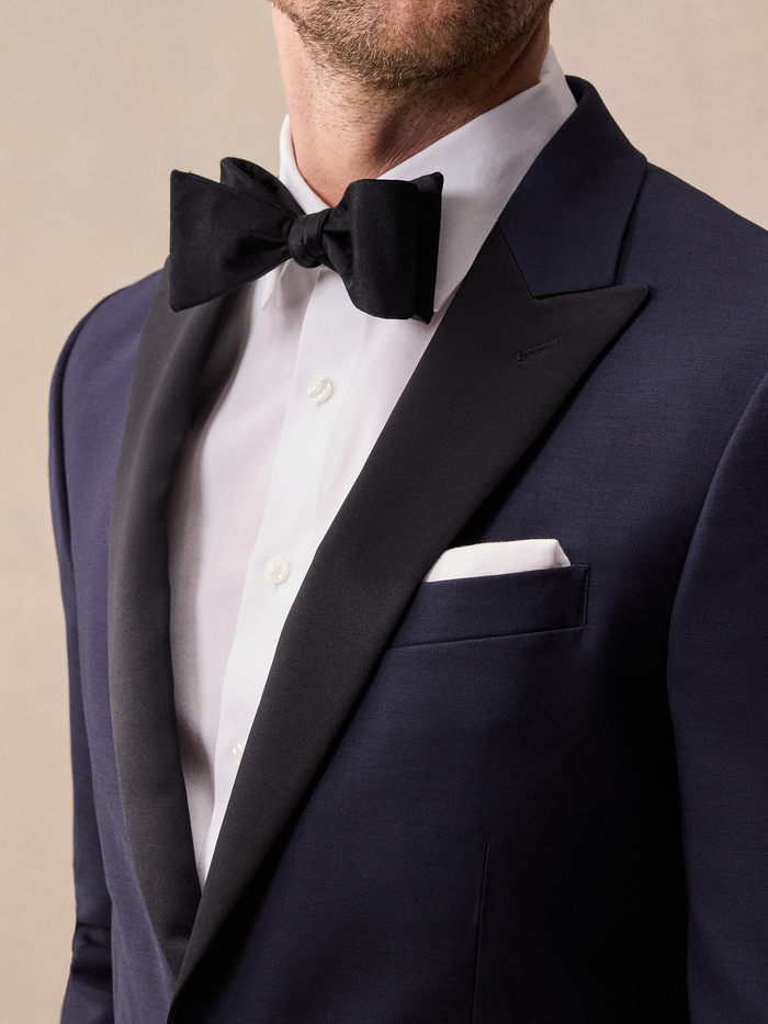 Close-up of a man in a midnight blue tuxedo with black satin peak lapel, black bow tie, and white pocket square.