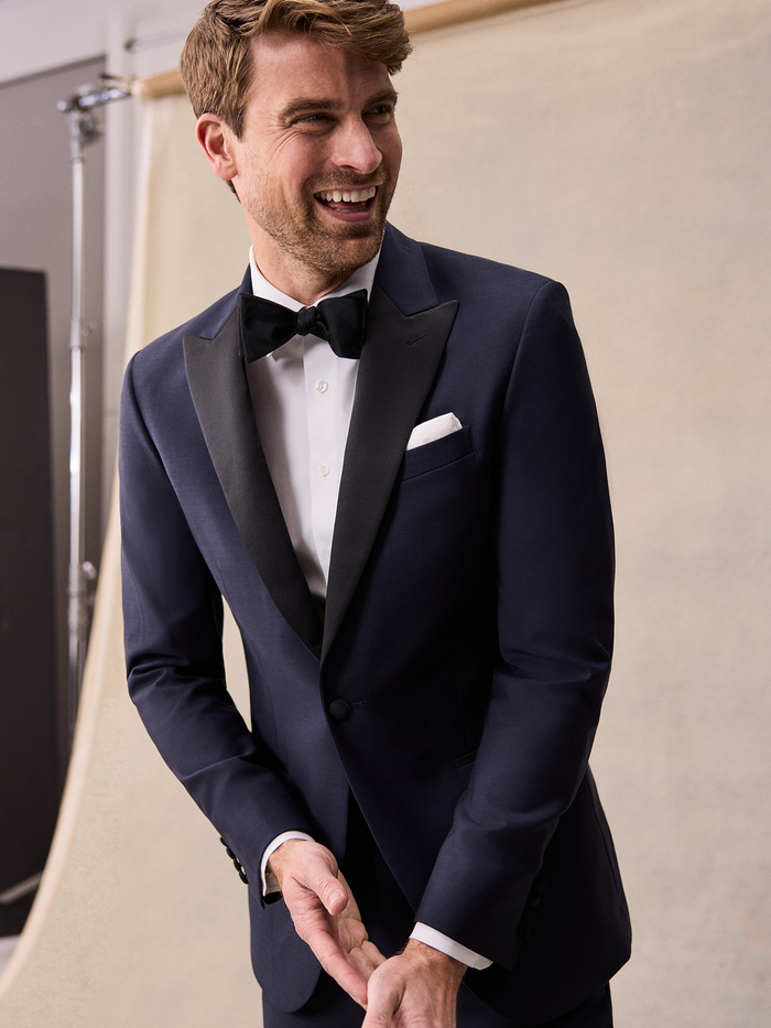 A smiling man in a midnight blue tuxedo with black satin peak lapels, black bow tie, and white pocket square.