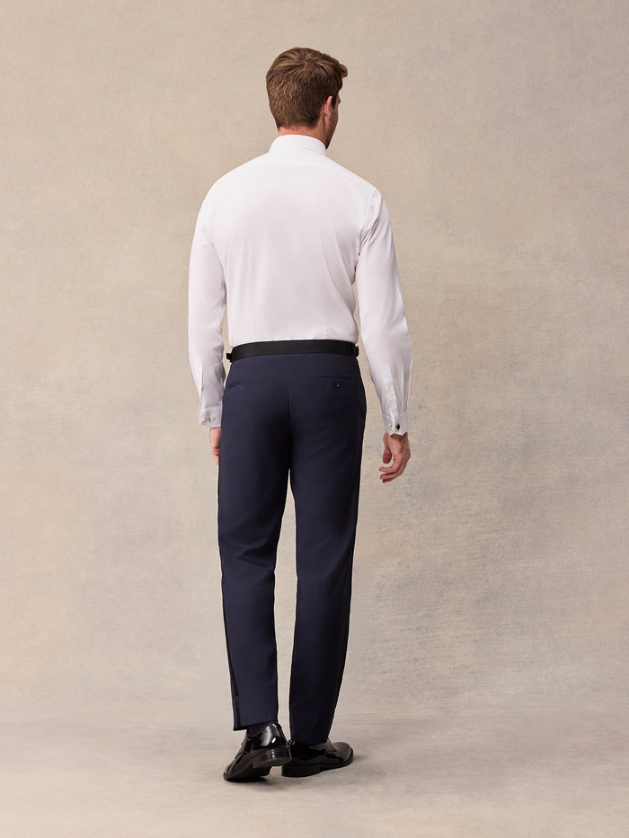 Back view of man in white tuxedo shirt with black waist tab and midnight blue tuxedo pants, full length
