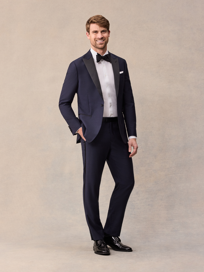 Man in midnight blue tuxedo with black satin peak lapels and black bow tie, full length, beige backdrop