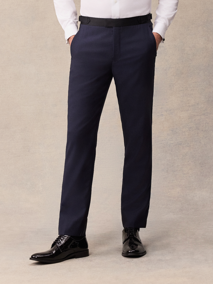Man in midnight blue tuxedo pants with black satin waistband tab, hands in pockets, front view