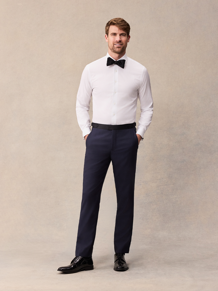 A man in a white tuxedo shirt with a black bow tie and midnight blue tuxedo pants, hands in pockets, full length.