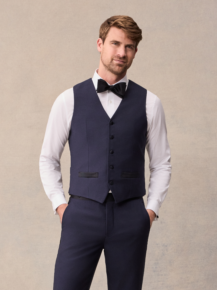 Man in midnight blue tuxedo vest over white shirt with black bow tie, hands in pockets, full length