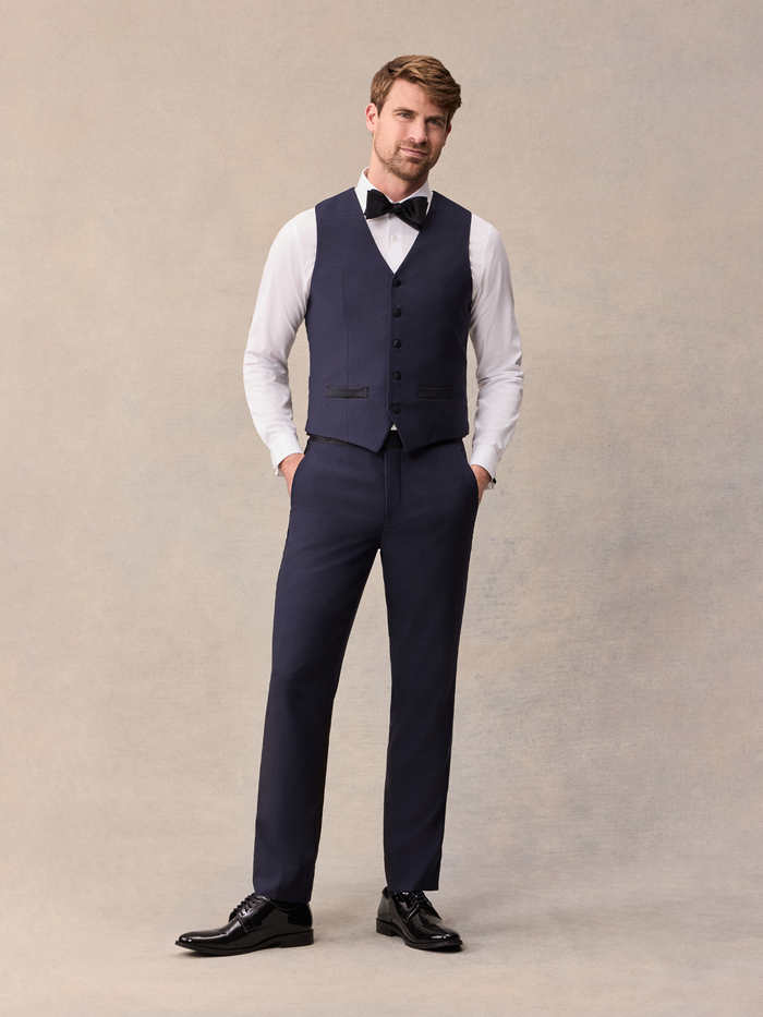 Man in midnight blue tuxedo vest over white shirt with black bow tie, hands in pockets, full length