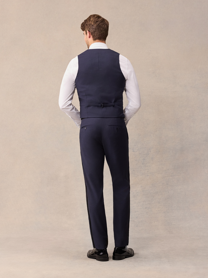 Back view of man in midnight blue tuxedo vest with satin strap and buckle over white shirt, full length
