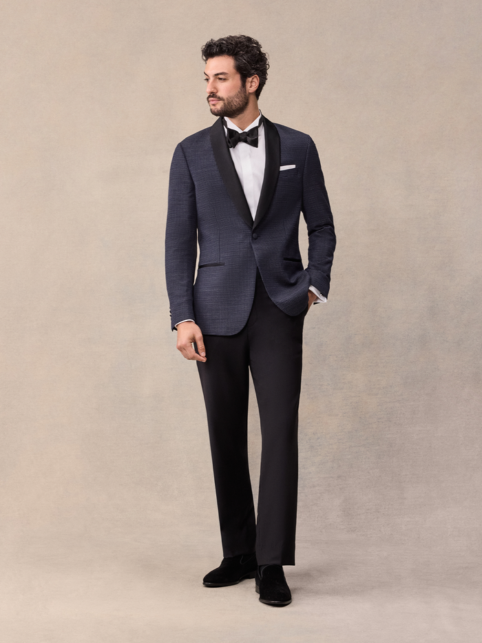 Man in midnight pin-dot tuxedo with black satin shawl lapel and bow tie, full length, tan backdrop