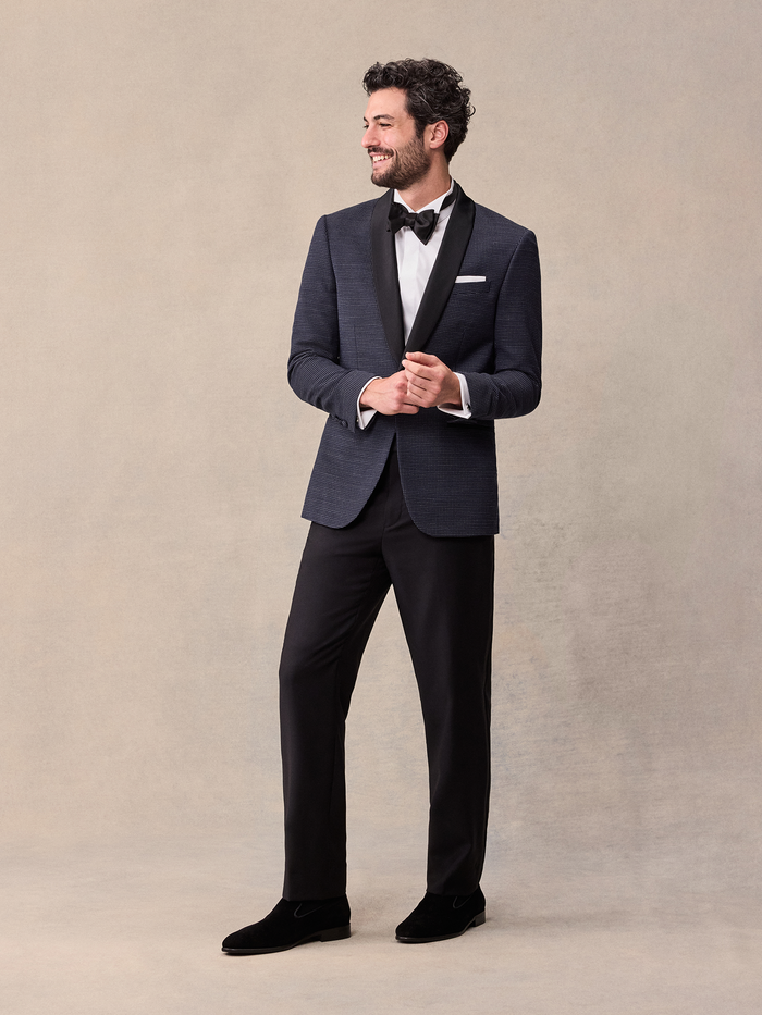 Man in midnight pin-dot tuxedo with black satin shawl lapel, hands at button, full length, tan backdrop