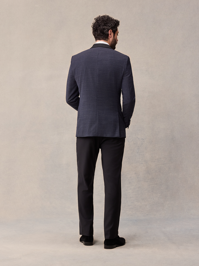 Back view of man in midnight pin-dot tuxedo jacket and black trousers, full length, tan backdrop