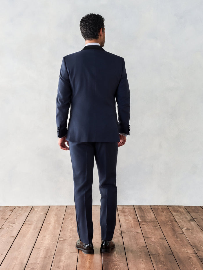 Back view of man in midnight shawl-lapel tuxedo jacket and trousers, full length on wood floor