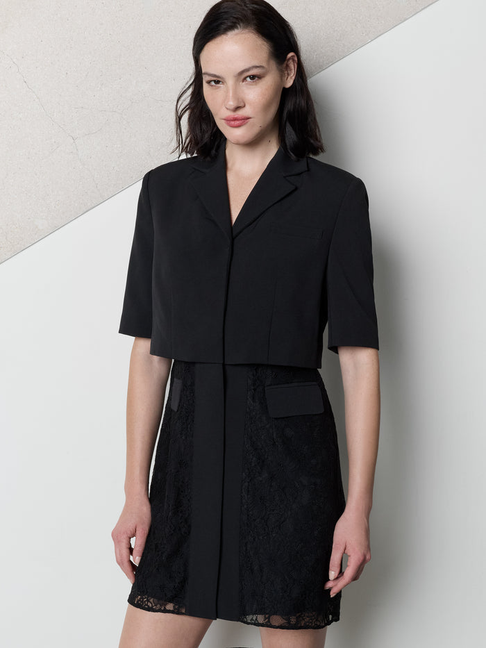 A woman wearing a black Dress with padded shoulders, short sleeves, lapel collars, V-neckline, front button fastening, a lace skirt and a tailored mid length fit.