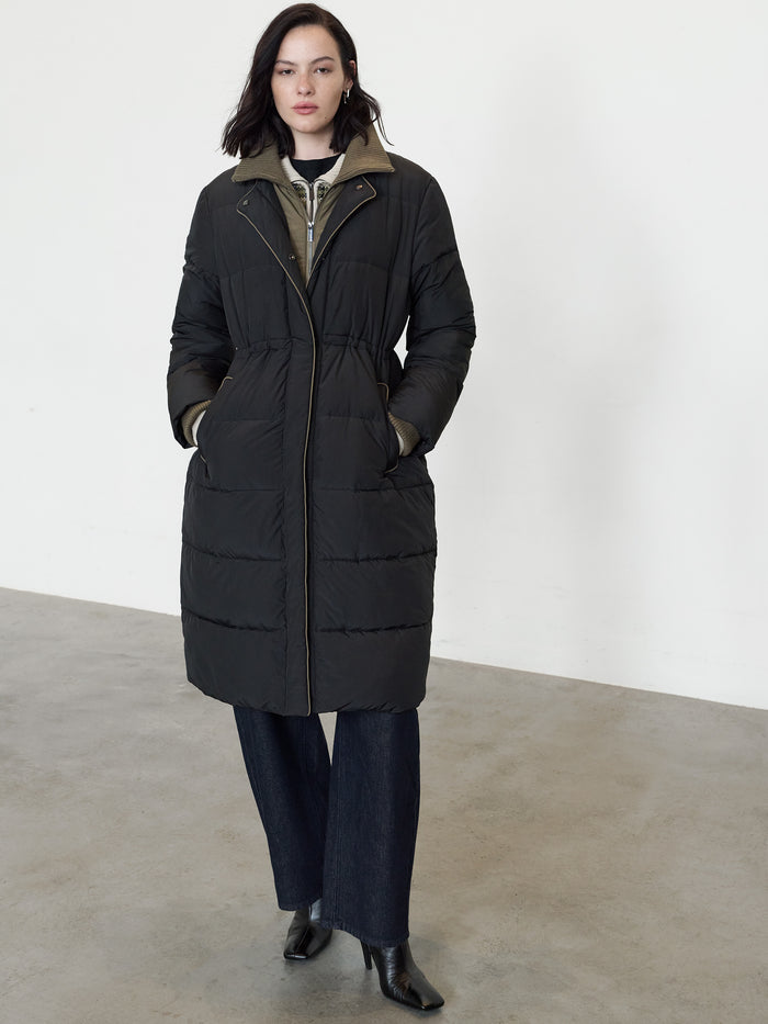 A woman wearing a black long-line puffer coat with a funnel neckline, long sleeves, concealed zip and popper fastening and 2 side pockets.