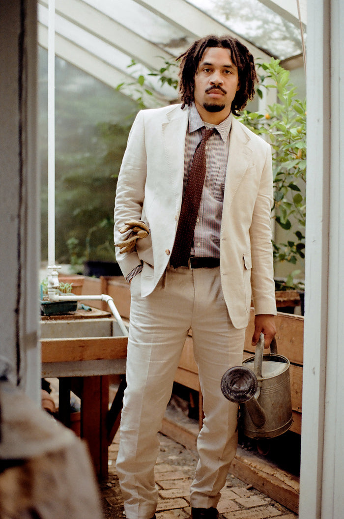 Man in mojave tan linen suit holding gardening gloves and a watering can, greenhouse doorway