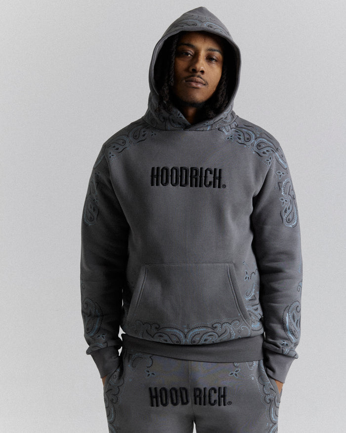 A man wearing a dark gray hooded sweatshirt and matching sweatpants with 'HOODRICH.' printed across the chest and upper thighs, featuring tonal paisley patterns on the sleeves and lower hem, against a light gray studio background.