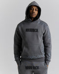 A man wearing a dark gray hooded sweatshirt and matching sweatpants with 'HOODRICH.' printed across the chest and upper thighs, featuring tonal paisley patterns on the sleeves and lower hem, against a light gray studio background.